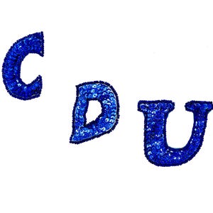 May include: Three blue sequined letters: C, D, and U, against a white background. The letters are in a bold, rounded font, and each is densely covered in small, shiny blue sequins. The sequins catch the light, creating a sparkling effect.