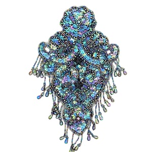 May include: A detailed, ornate, beaded applique in shades of blue, teal, and purple. The design features a central, symmetrical pattern with flowing lines and teardrop-shaped bead fringe. The sequins and beads create a shimmering effect against the white background.