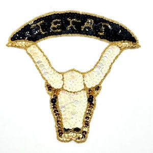 May include: A white and gold sequined bull's head with black eyes and a black sequined banner above that reads "TEXAS".
