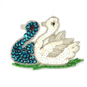 May include: A decorative patch featuring two swans, one covered in iridescent blue sequins and the other in white sequins, with clear glass beads and a small black bead for the eye. The swans rest on a base of bright green cylindrical glass beads.