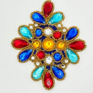 May include: A colorful, ornate embellishment with a symmetrical design. It features teardrop-shaped and round gemstones in red, blue, turquoise, yellow, and clear, surrounded by gold-colored beads. The piece is set against a white background.