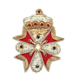 May include: An ornate, embroidered brooch featuring a crown design atop a red and gold cross-shaped emblem. The brooch is embellished with silver thread, red and blue gemstones, and a central pearl. The intricate details suggest a vintage or regal aesthetic.