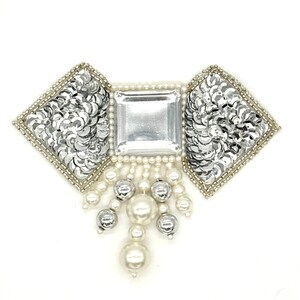 May include: A decorative silver and pearl embellishment. The design features a central square crystal, flanked by two sequined diamond shapes. Pearl beads and silver orbs hang below, creating a glamorous, elegant look.