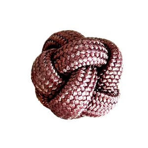 May include: A close-up of a decorative knot made of woven, reddish-brown material. The knot is tightly woven, with a textured surface and a rounded shape. The background is plain white.