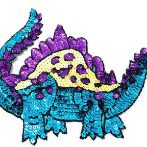 May include: A sequined dinosaur patch in shades of turquoise, purple, and yellow. The dinosaur has a long tail and a spiky back, with black beaded outlines. The sequins create a shimmering effect, perfect for embellishing clothing or accessories.