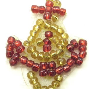 May include: A beaded ornament in a starburst design. The ornament features a mix of red and gold beads, arranged on a white, textured backing. The beads are small and cylindrical, creating a textured surface. The overall shape is symmetrical and decorative.