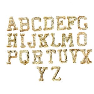 May include: Gold sequin alphabet letters, A through Z, arranged on a white background. Each letter is made of small, shiny gold sequins, creating a textured and decorative appearance. The letters are in a serif font.