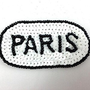 May include: A white sequined patch with the word "PARIS" spelled out in black beads. The patch has a black beaded border and is oval-shaped.