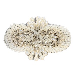 May include: An oval-shaped, cream-coloured beaded embellishment featuring clear rhinestones and faux pearls. The piece is densely covered in small, white beads, creating a textured surface. The design is symmetrical and ornate, suitable for clothing or accessories.