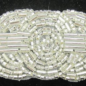 May include: A silver beaded embellishment with a central circular design and two horizontal rows of beads. The embellishment is on a white mesh backing.