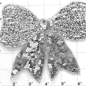 May include: A silver sequined bow applique measuring approximately 6 inches wide and 4 inches tall. The bow is made of small, round sequins sewn together in a layered pattern.