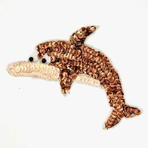 May include: A dolphin-shaped patch crafted with metallic bronze sequins covering the body and light champagne-colored sequins on the underbelly. The design features two small black beads for eyes and is outlined with a border of tiny white seed beads.