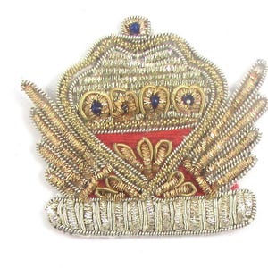 May include: A gold embroidered patch with a crown and wings. The crown has red and gold details and blue jewels. The wings are gold and have a red center.
