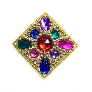 May include: A square-shaped, gold-toned brooch with a central red gemstone surrounded by an array of colorful, faceted gems. The brooch features blue, purple, green, and pink stones, and is bordered by small, gold-colored beads.
