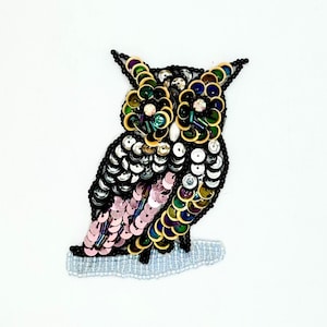May include: An owl-shaped patch crafted with black, silver, pink, and iridescent green sequins and beads. The owl features large circular eyes, a layered feather design, and is perched on a horizontal bar made of small, clear, light-reflecting beads.