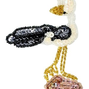 May include: A decorative patch shaped like a bird, crafted with white, black, and iridescent sequins. The bird features a gold beaded beak, long gold beaded legs, and stands upon a cluster of light pink sequins. The design is detailed with beads.