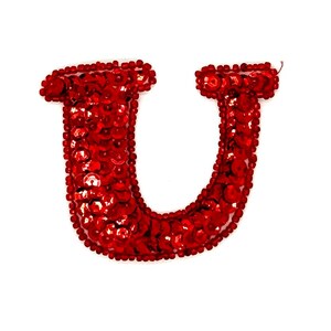 May include: A red sequined letter "U" against a white background. The letter is densely covered in small, shiny red sequins, giving it a textured appearance. The edges are outlined with small red beads.