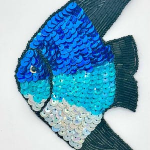 May include: A fish-shaped patch featuring a gradient of blue, turquoise, and iridescent white sequins on the body. The fins and tail are outlined and filled with dark navy blue glass beads, creating a distinct contrast against the sequined body.