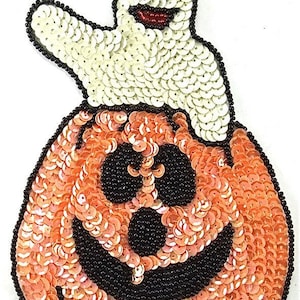 May include: A Halloween-themed sequined patch featuring a smiling jack-o'-lantern with a friendly ghost on top. The pumpkin is orange with black facial features and the ghost is white. The patch is outlined with black beads.