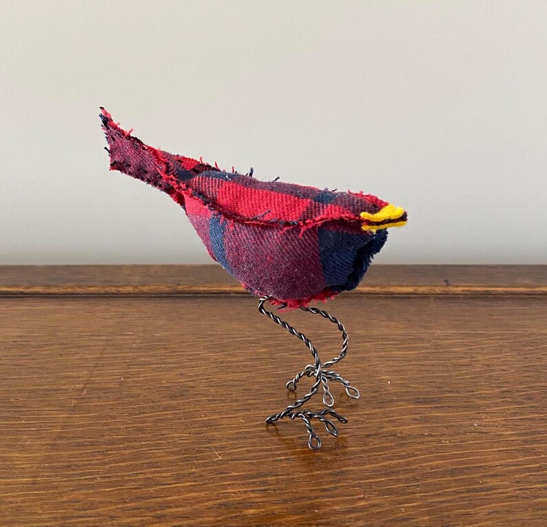 May include: A small, red and blue plaid fabric bird with a yellow beak, standing on a black wire base.