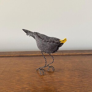 K&ouml;nnte beinhalten: Ein kleiner Vogel mit schwarz-wei&szlig;em Muster, gelbem Schnabel und Beinen aus Draht. Der Vogel steht auf einer Holzoberfl&auml;che.