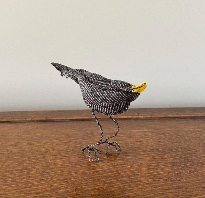 K&ouml;nnte beinhalten: Ein kleiner Vogel mit schwarz-wei&szlig;em Muster, gelbem Schnabel und Beinen aus Draht. Der Vogel steht auf einer Holzoberfl&auml;che.