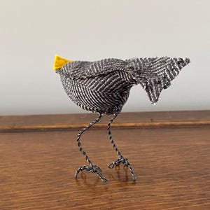 K&ouml;nnte beinhalten: Ein kleiner grauer und wei&szlig;er Vogel mit einem gelben Schnabel aus Stoff und Drahtbeinen. Der Vogel steht auf einer Holzoberfl&auml;che.