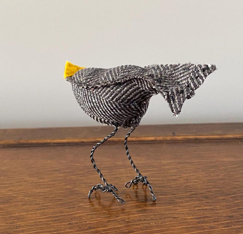 K&ouml;nnte beinhalten: Ein kleiner grauer und wei&szlig;er Vogel mit einem gelben Schnabel aus Stoff und Drahtbeinen. Der Vogel steht auf einer Holzoberfl&auml;che.