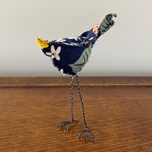 May include: A small, blue and green fabric bird with a yellow tail, standing on black wire legs. The bird is handmade and has a whimsical, folk art style.