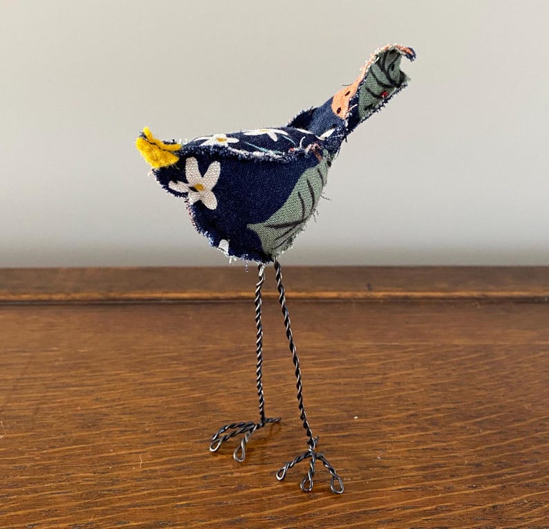 May include: A small, blue and green fabric bird with a yellow tail, standing on black wire legs. The bird is handmade and has a whimsical, folk art style.