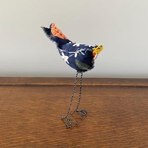 May include: A small, handmade bird figurine with a blue and white floral fabric body, orange and black fabric tail, yellow beak, and wire legs.