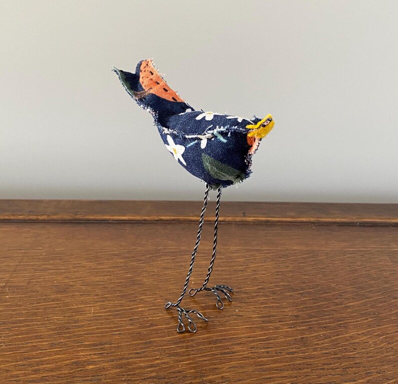 May include: A small, handmade bird figurine with a blue and white floral fabric body, orange and black fabric tail, yellow beak, and wire legs.