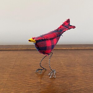 May include: A small, handmade bird figurine made from red and black checked fabric. The bird has wire legs and a yellow beak.