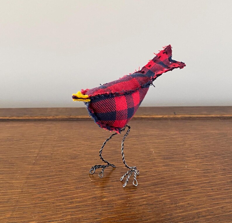 May include: A small, handmade bird figurine made from red and black checked fabric. The bird has wire legs and a yellow beak.