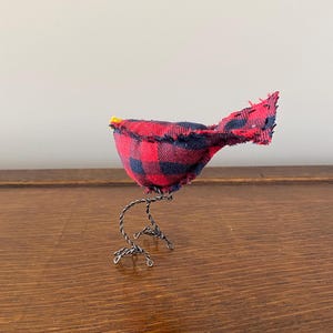 May include: A small, handmade bird figurine made from red and black checked fabric. The bird has wire legs and a yellow beak.