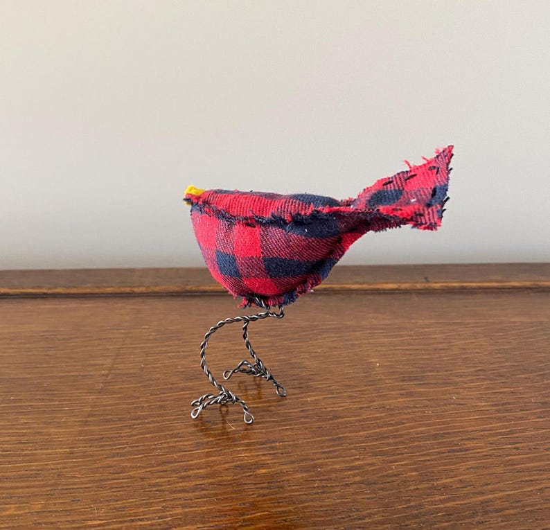 May include: A small, handmade bird figurine made from red and black checked fabric. The bird has wire legs and a yellow beak.