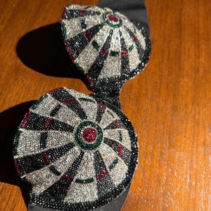 May include: A black bra with dartboard-style embellishments. The cups are covered in silver, black, red, and green beads, with a black satin band. The design resembles a dartboard.