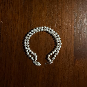 May include: A double-strand pearl bracelet with a silver clasp. The bracelet features small, round, white pearls. The clasp is a decorative oval shape with a secure closure. The bracelet is displayed on a dark brown wooden surface.