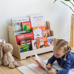May include: A wooden bookshelf with multiple shelves filled with colorful children's books. The bookshelf is in a room with a rug and a child reading a book on the floor.