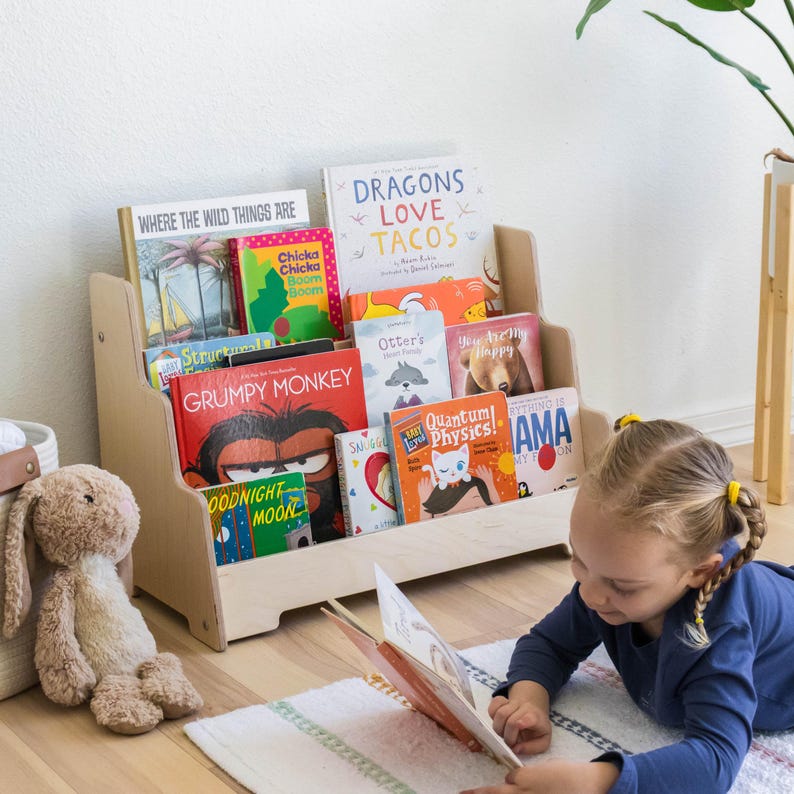 May include: A wooden bookshelf with multiple shelves filled with colorful children's books. The bookshelf is in a room with a rug and a child reading a book on the floor.