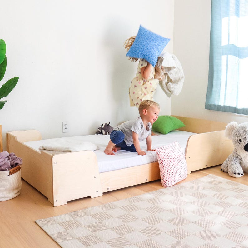 May include: A light-coloured wooden toddler bed with a white mattress. A child is climbing on the bed. A green cushion, a red and white polka dot cushion, and a stuffed koala are nearby. A patterned rug is on the floor.