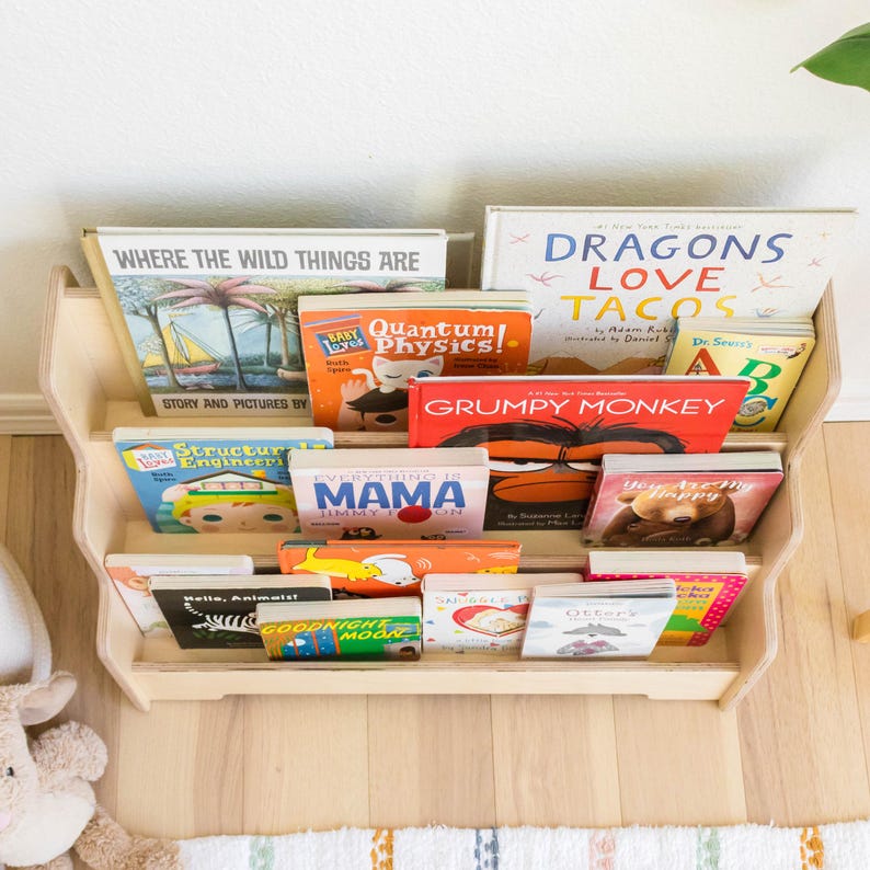 May include: A wooden bookshelf with five shelves filled with colorful children's books. The books have titles such as "Where the Wild Things Are", "Dragons Love Tacos", "Grumpy Monkey", "Everything is Mama", and "Goodnight Moon".
