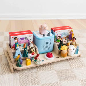 May include: A wooden toy organizer with a light blue speaker, a pink pig figurine, and various character figurines. Two red boxes with white text are also present. The organizer is on a patterned rug.