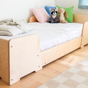 May include: A light-coloured wooden bed frame with a white mattress, pillows, and soft toys. The bed has a simple design with rounded corners and visible screws. A folded blanket and a patterned rug are also visible.