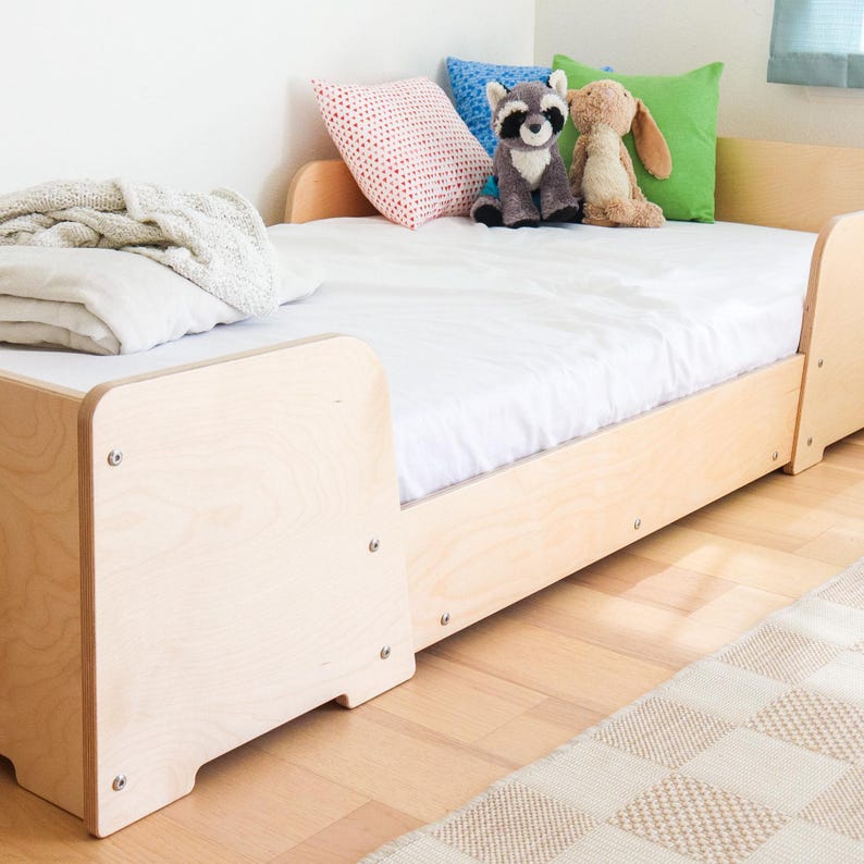 May include: A light-coloured wooden bed frame with a white mattress, pillows, and soft toys. The bed has a simple design with rounded corners and visible screws. A folded blanket and a patterned rug are also visible.