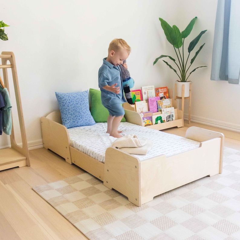 May include: A light wood toddler bed with a patterned mattress, a blue cushion, and a green cushion. A child in blue clothing is standing on the bed. A small bookshelf with books is in the background. A beige and white checkered rug is on the floor.