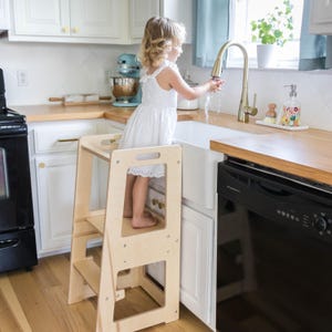 May include: A light wood step stool with two steps and a safety rail. A young child is standing on the top step, reaching for a sink.
