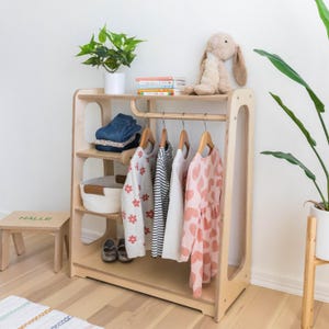 K&ouml;nnte beinhalten: Ein Kinderkleiderschrank aus Holz mit Regalen, einer Kleiderstange und einem kleinen Hocker. Der Schrank ist aus hellem Holz und hat eine nat&uuml;rliche Oberfl&auml;che. An der Stange h&auml;ngen Kleider und auf einem der Regale steht ein Korb. Der Hocker ist mit "Halle" beschriftet.