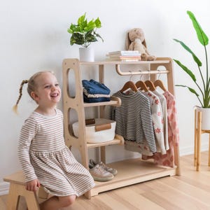 K&ouml;nnte beinhalten: Ein helles Kinderkleiderschrank aus Holz mit einer Kleiderstange, zwei Regalen und einem unteren Regal. Ein Kind sitzt auf einem Holzhocker vor dem Schrank. Der Schrank hat einen wei&szlig;en Korb auf dem oberen Regal und einen braunen Ledergriff.