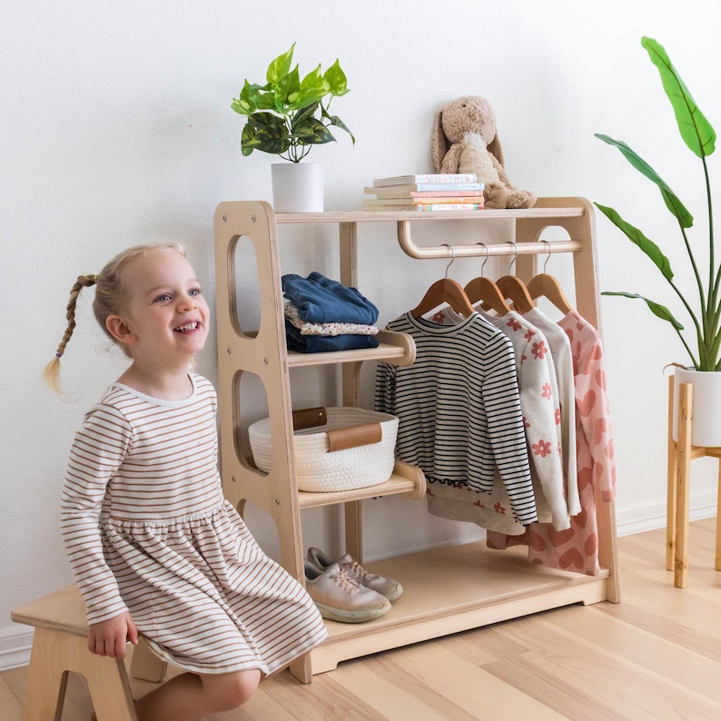 K&ouml;nnte beinhalten: Ein helles Kinderkleiderschrank aus Holz mit einer Kleiderstange, zwei Regalen und einem unteren Regal. Ein Kind sitzt auf einem Holzhocker vor dem Schrank. Der Schrank hat einen wei&szlig;en Korb auf dem oberen Regal und einen braunen Ledergriff.